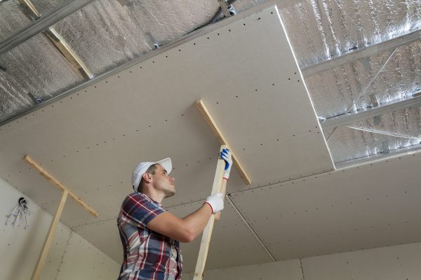 Young worker in protection work gloves fixing wooden holders for drywall suspended ceiling to metal frame insulated with shiny aluminum foil. DIY, do it yourself concept.